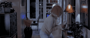 Drew Barrymore in white jeans and a cream jumper holding a phone in her home and then jumping before screaming.