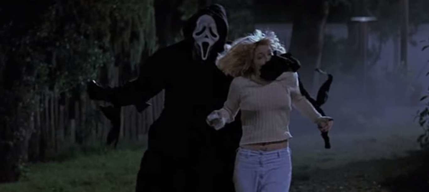 Drew Barrymore being held by the mouth by someone all in black with a scream mask on. Drew is wearing white jeans and a beige jumper.