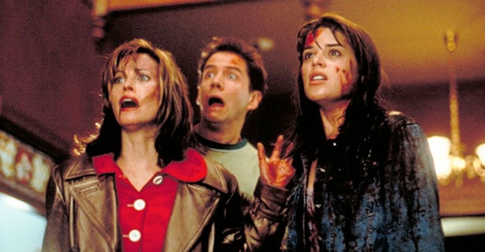 Three people stood together, one woman in a brown coat and red top, a guy with a grey t-shirt on and another woman in a jeans jacket. They are all covered in blood.