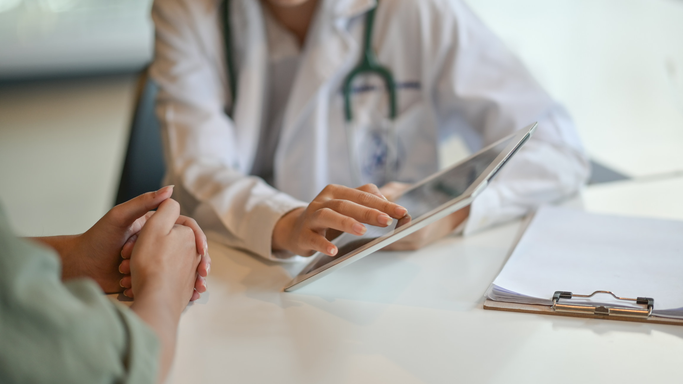 A stock image of a doctor tapping a tablet as they sit across from a patient at a table