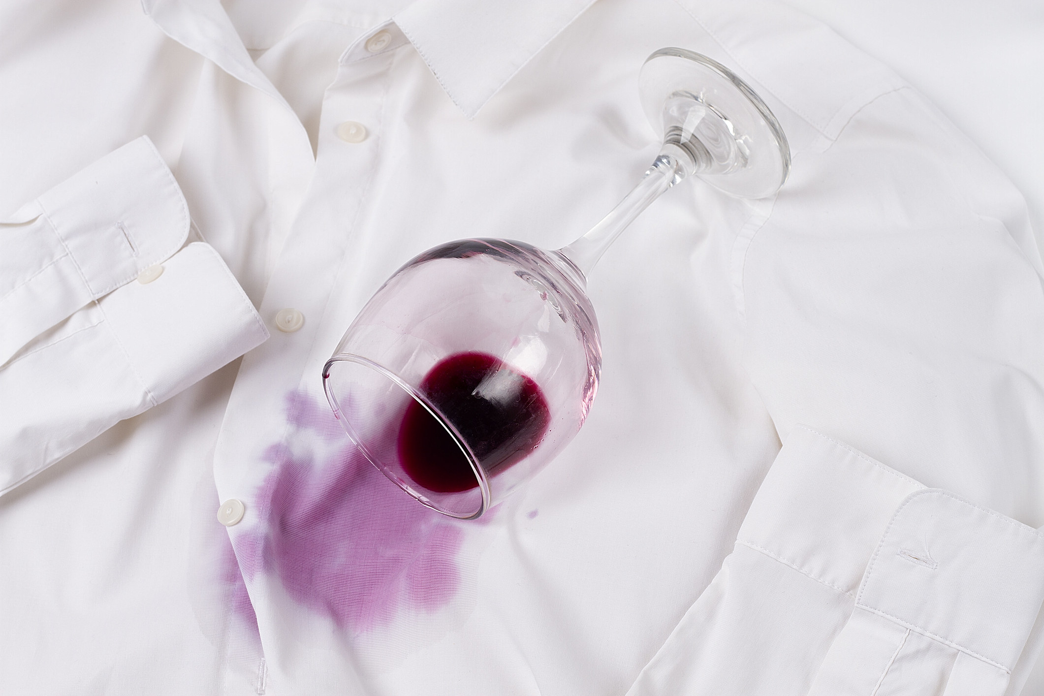 A stock image of a tipped over glass of red wine that is staining a white button down shirt
