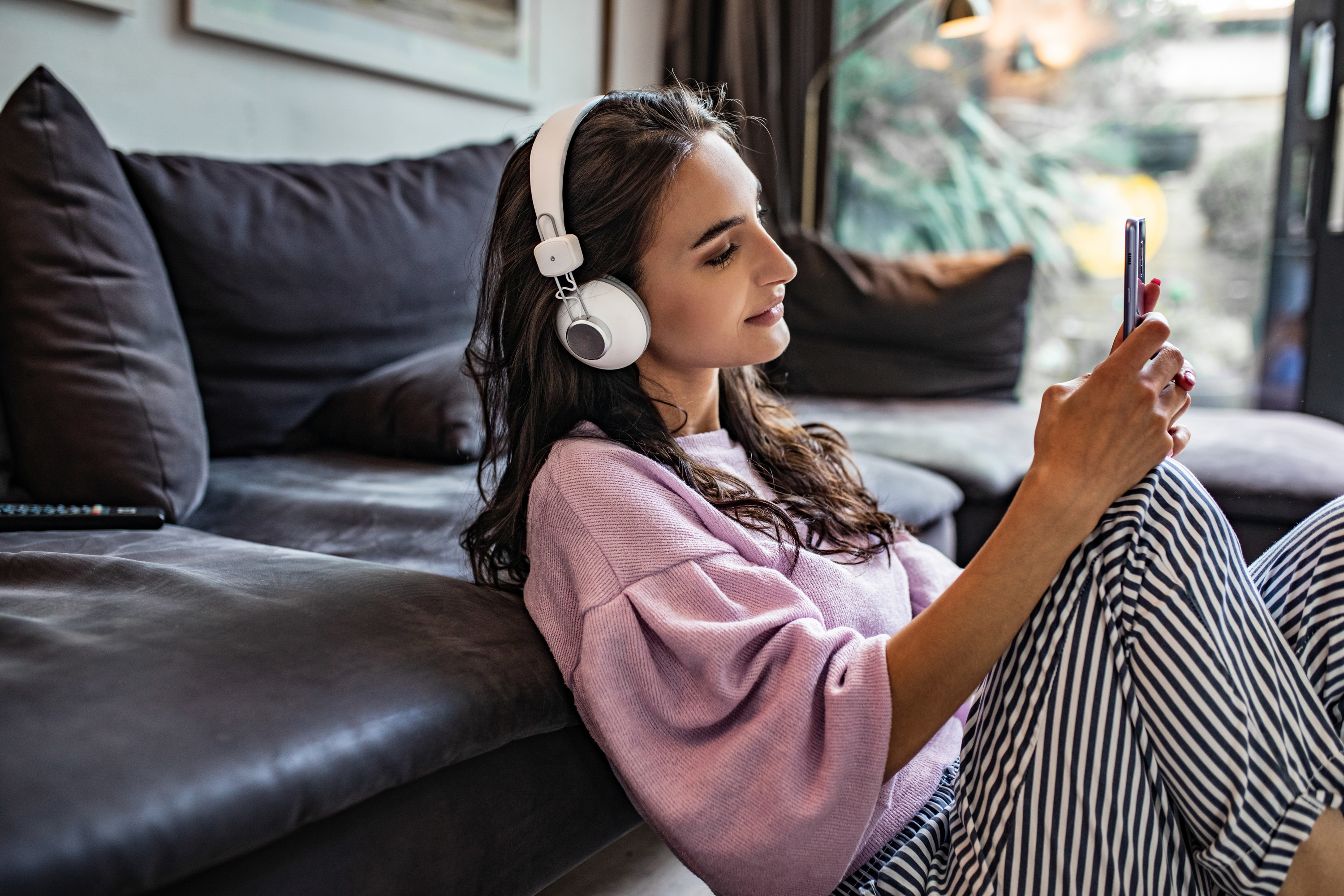 A girl in a pink sweatshirt sitting against a couch listens to a podcast through headphones