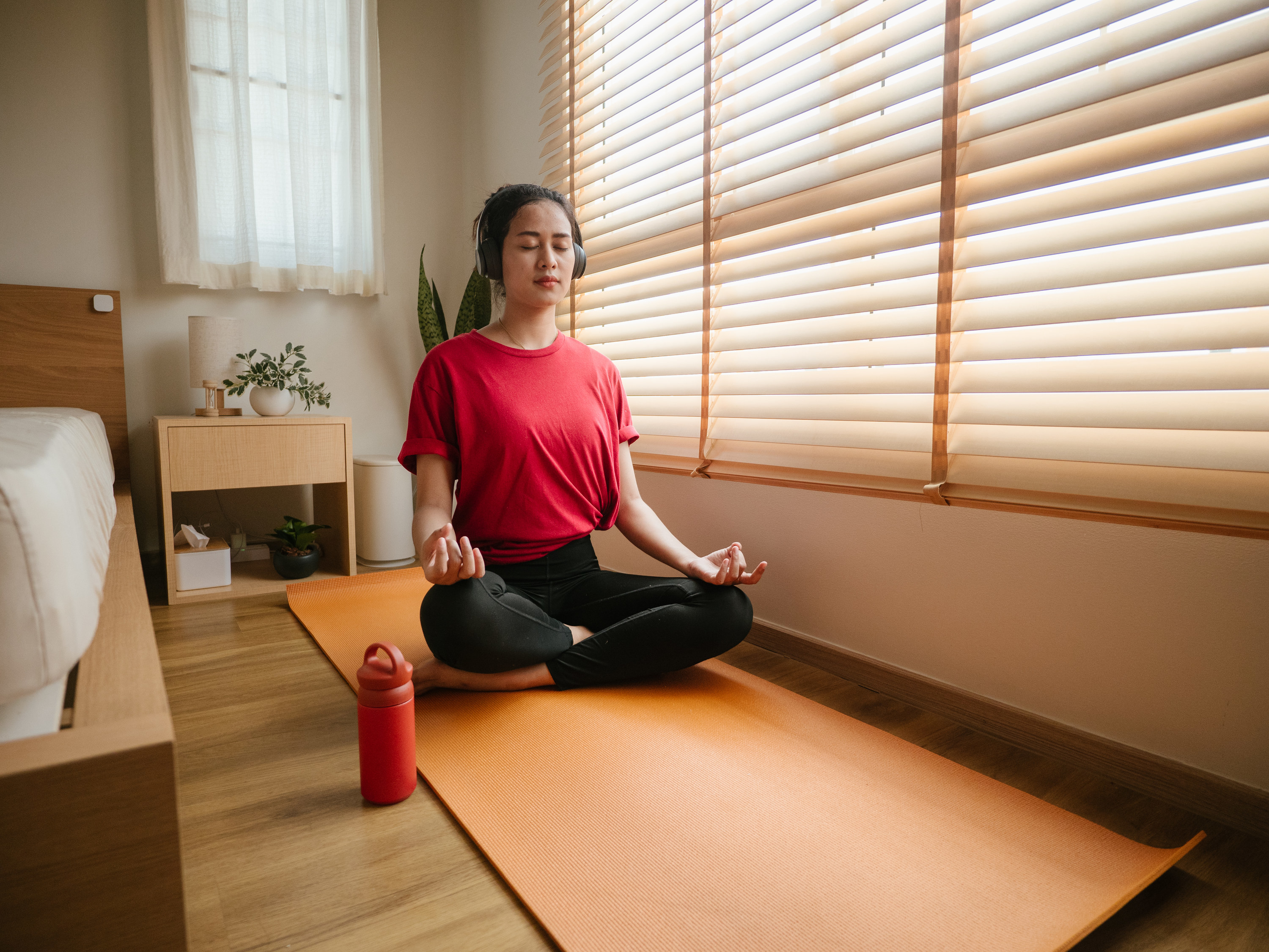 A woman in a red shirt sits meditating with her legs crossed and headphones on