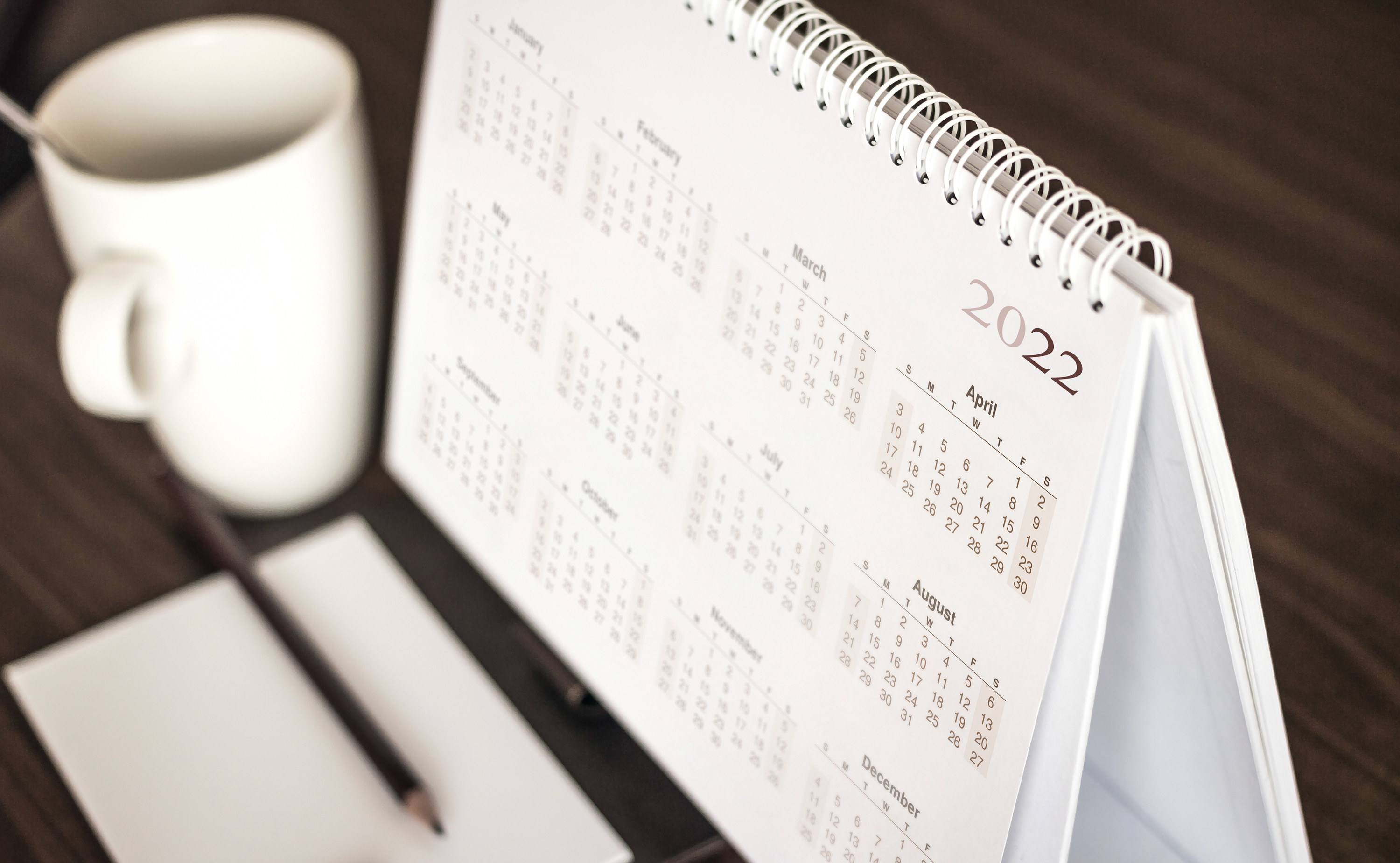 A notebook sized calendar propped up on a desk next to a mug