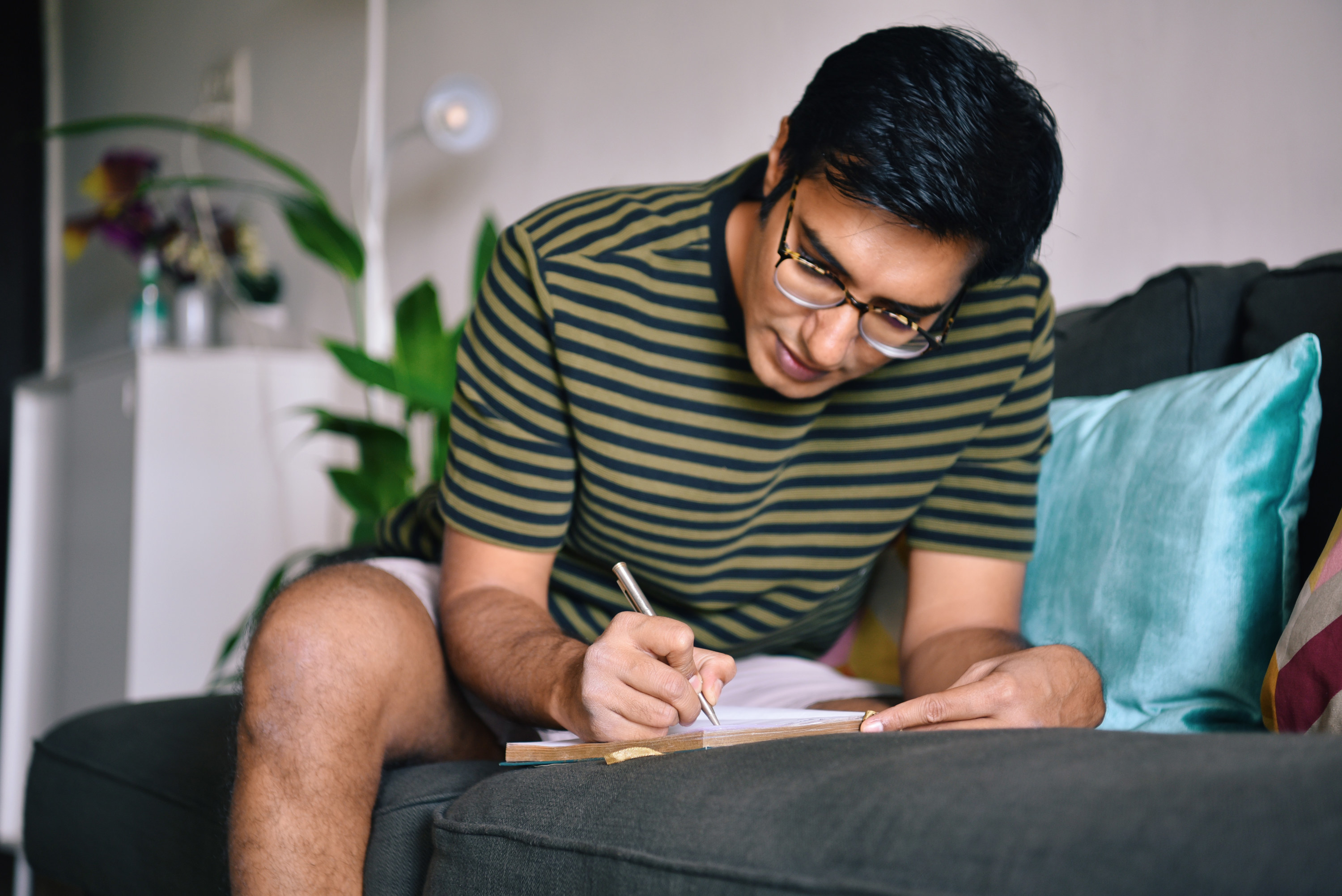 A young man in a green striped shirt writes in a journal on the couch