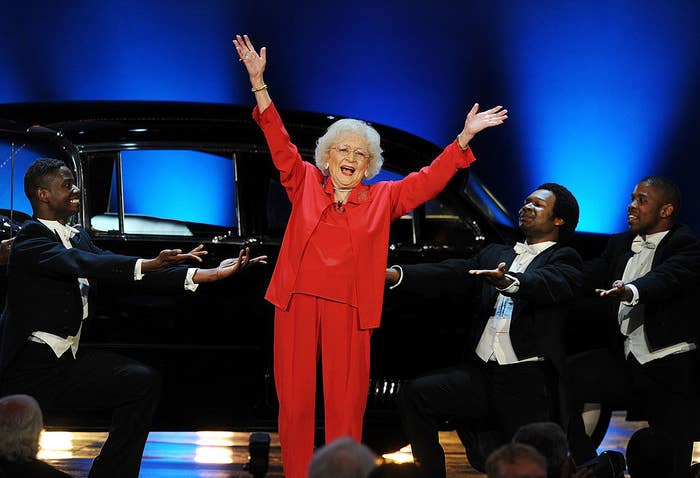 Betty White with her arms in the air onstage as she&#x27;s surrounded by background dancers in tuxedos