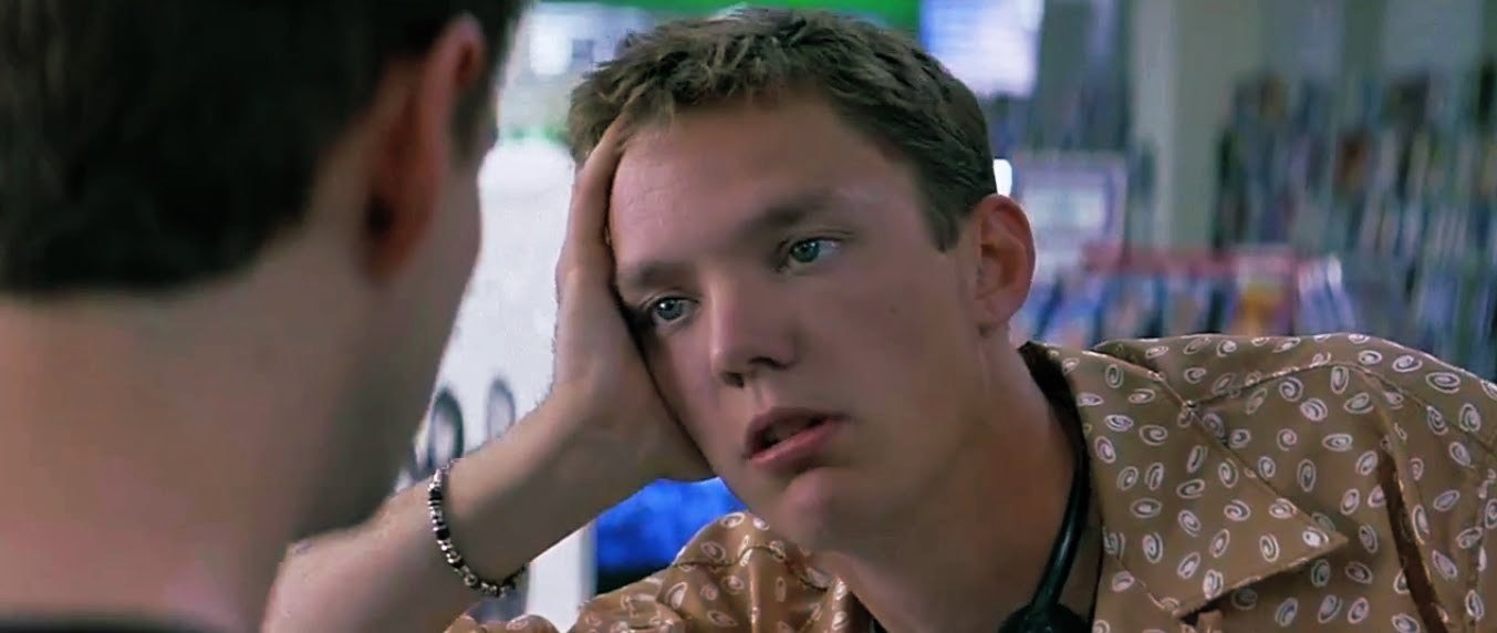 Matthew Lillard in a light brown shirt with a pattern on it, holding his head while he leans on something looking bored.