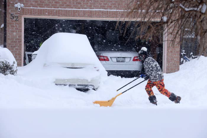 A resident cleans snow in front of a snow-covered car in a driveway.