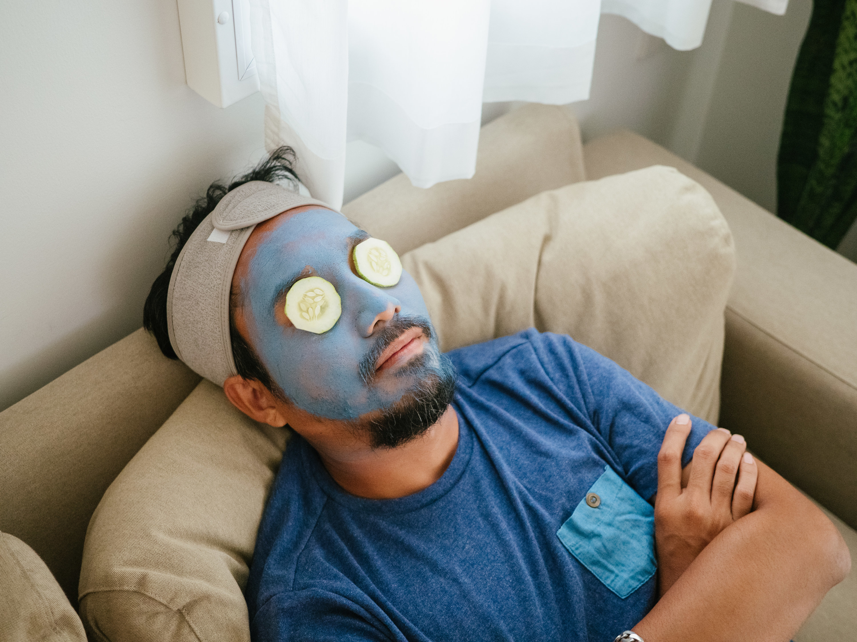 A man leans back with a blue clay face mask on and cucumber slices on his eyes