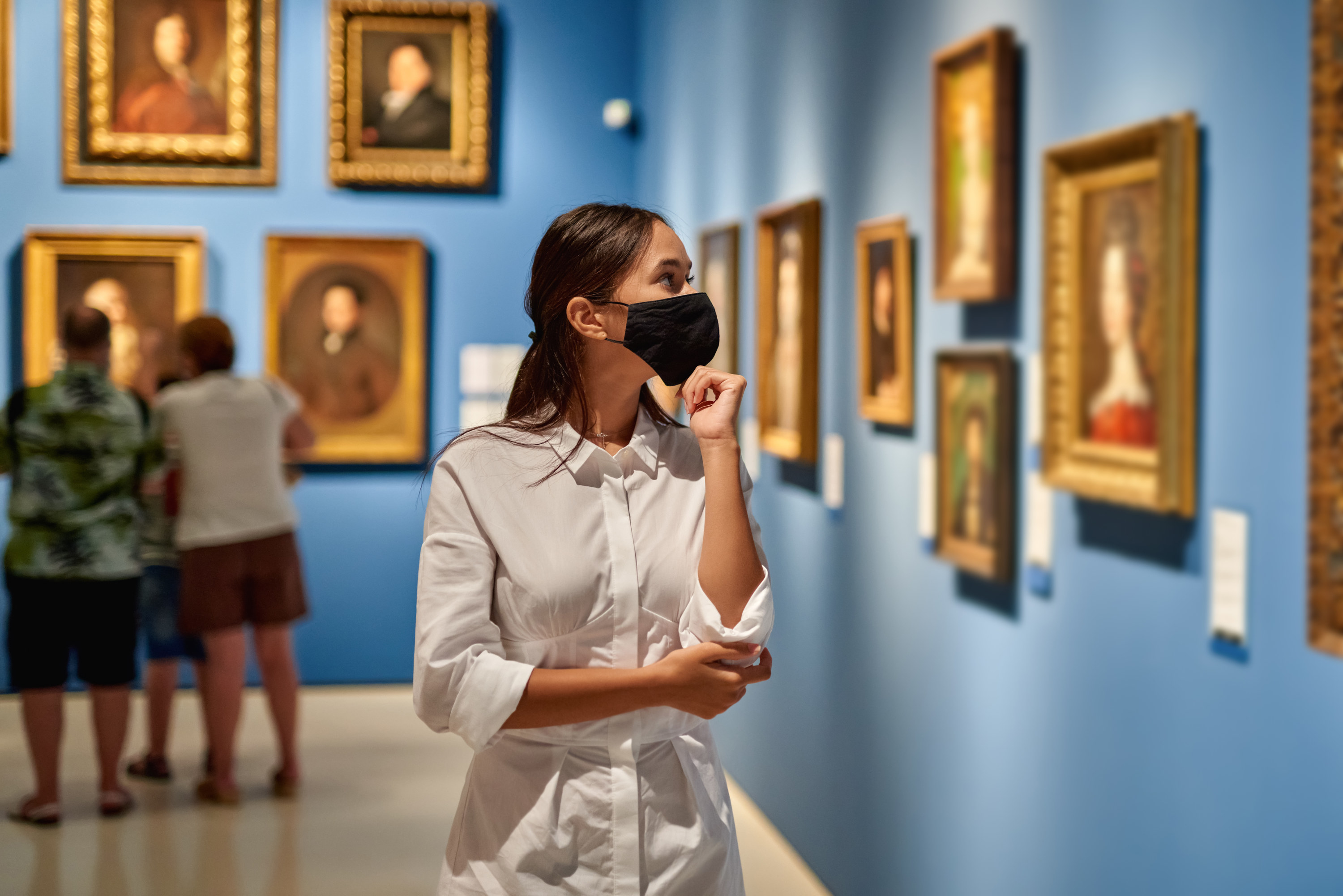 A woman in a white button-up and a black face masks takes in paintings at a museum
