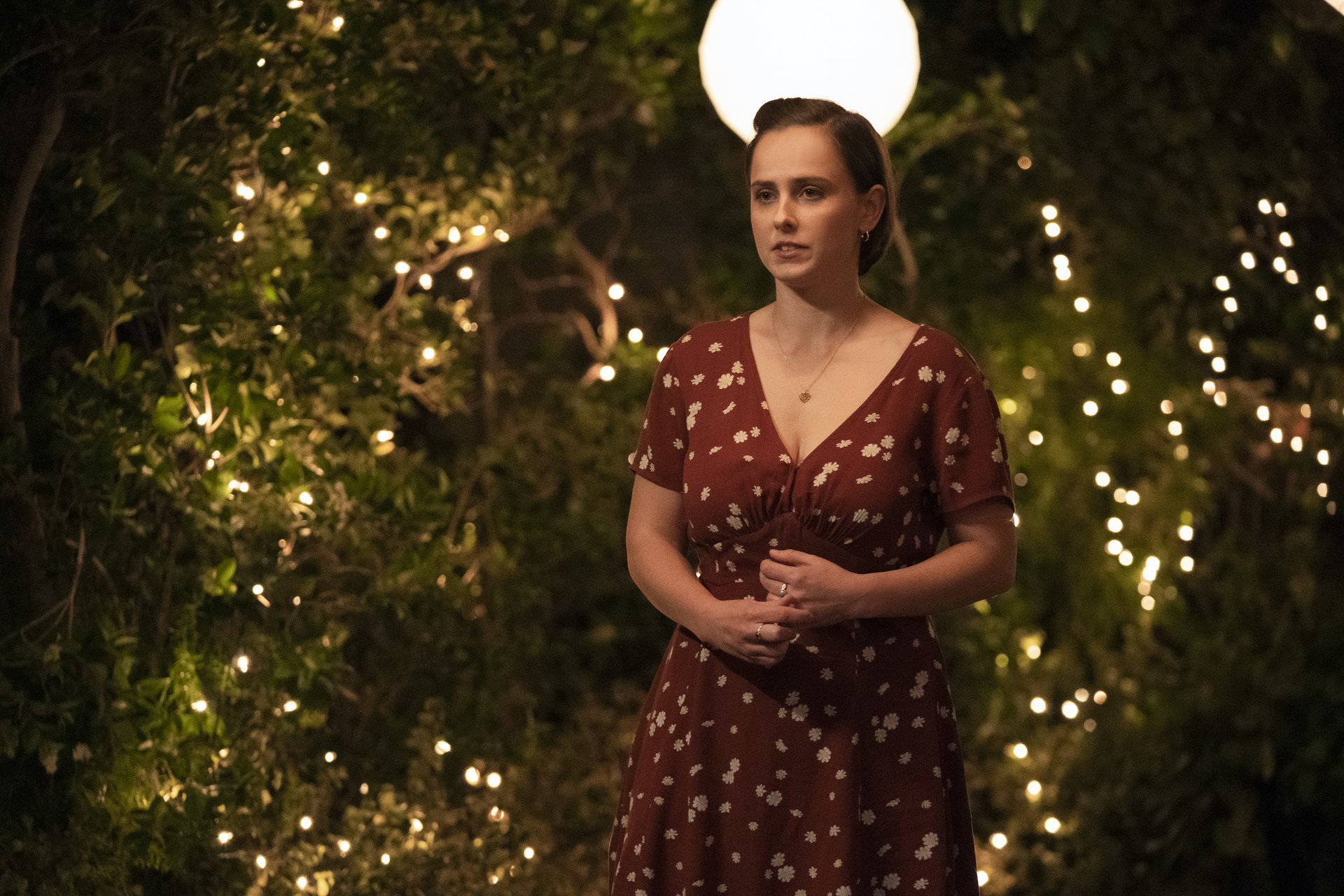 A white teenage girl in a red dress stands in front of bushes with lights in them