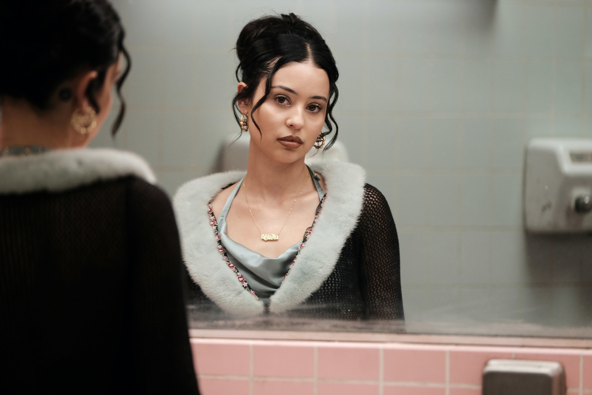 A teen girl with black hair looks at herself in a bathroom mirror