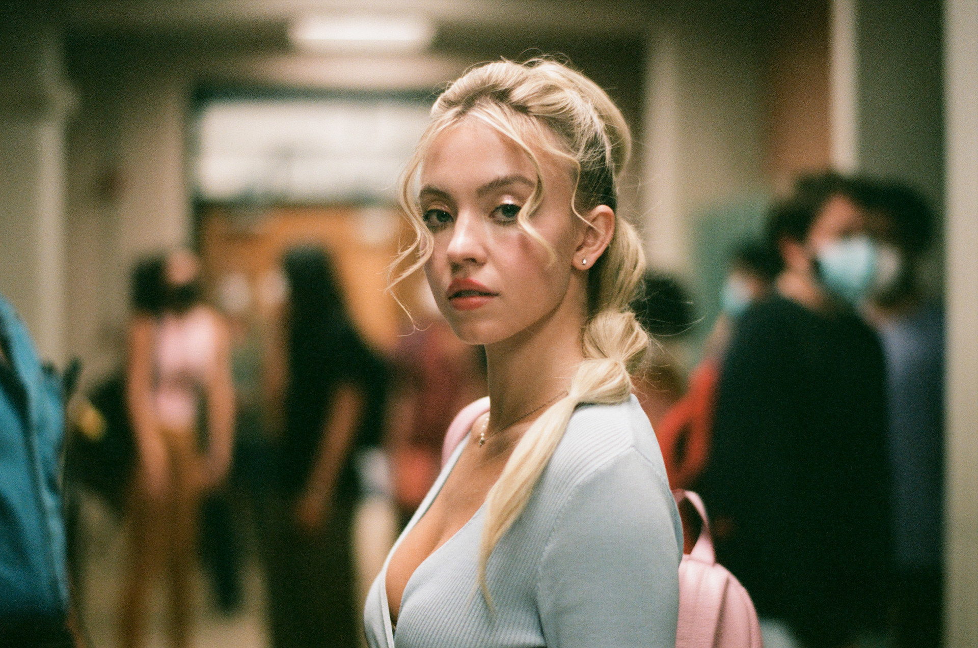 A blonde white teenage girl stands in a crowned high school hallway
