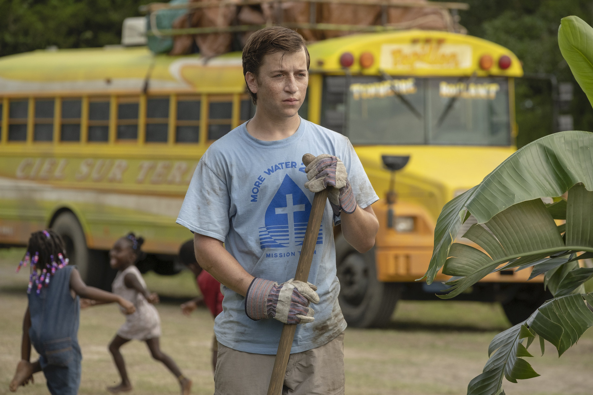 A white teen boy stands with work gloves in front of a bus.
