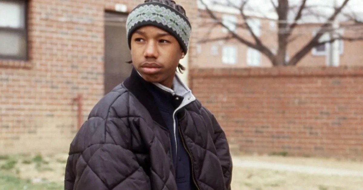A Black teenage boy stands outside wearing a hat and puffy black coat