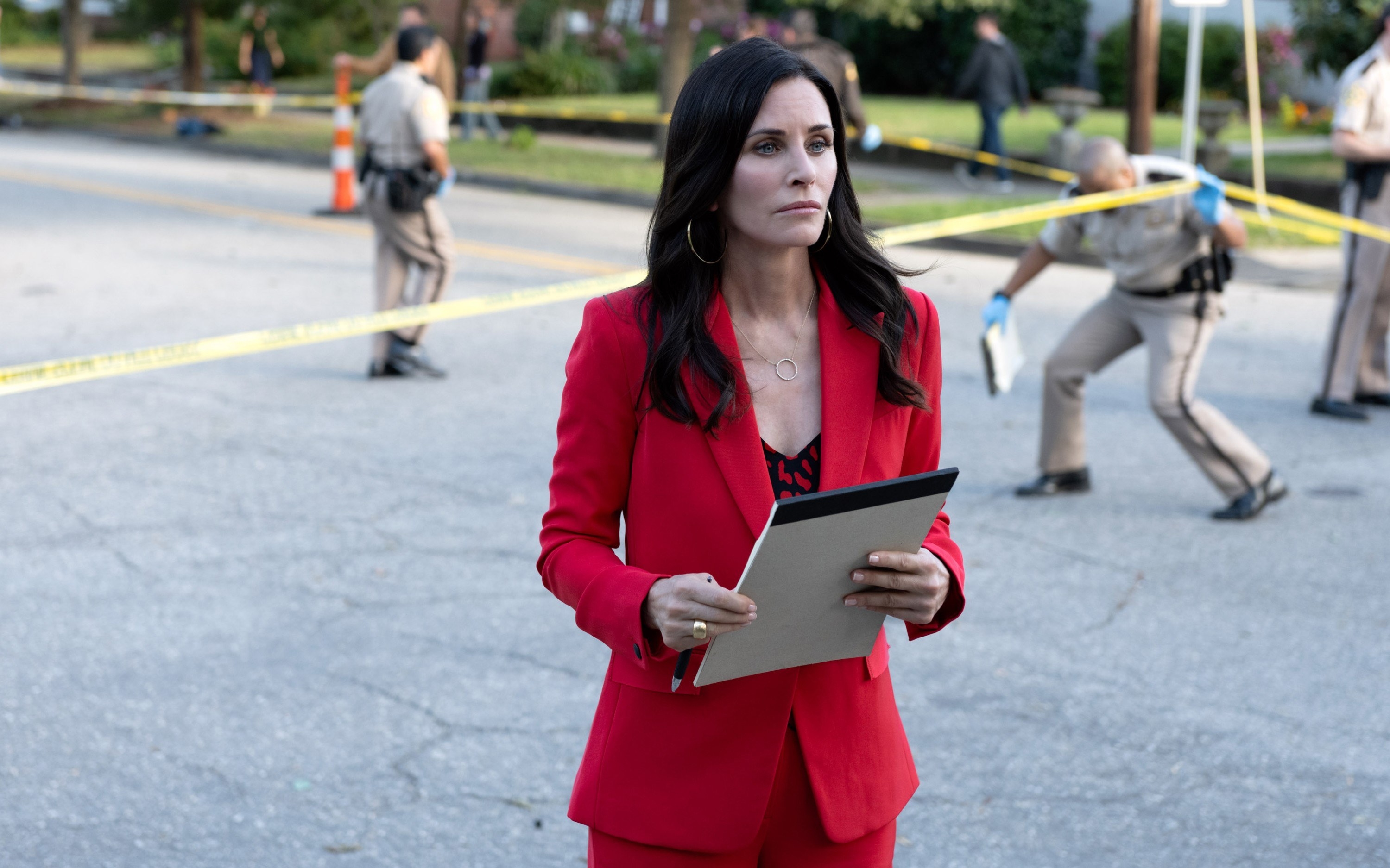Gale Weathers reporting for duty in a power suit outside a cordoned-off crime scene and holding a large notepad