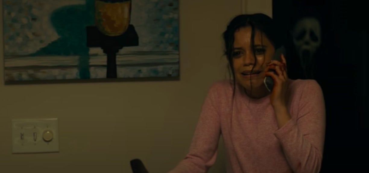 Tara holding a knife with a phone to her ear while Ghostface runs up behind her from the dark in &quot;Scream&quot; (2022)