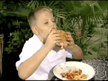 A young boy eating a burger and having ketchup and mustard spill out on his clean shirt