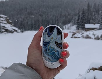 Reviewer is holding the white and blue marble hand warmer in their hand outside surrounded by snow