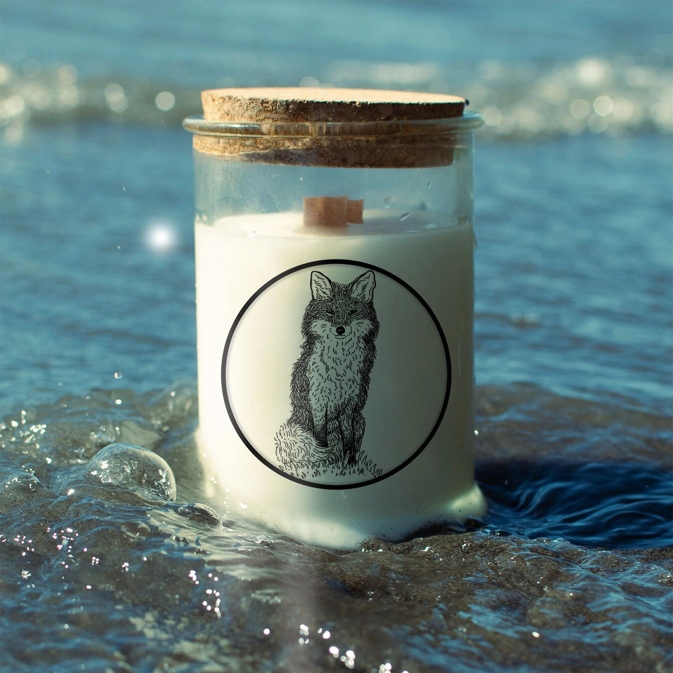 glass candle with cork top and fox illustration; it is placed in the ocean.