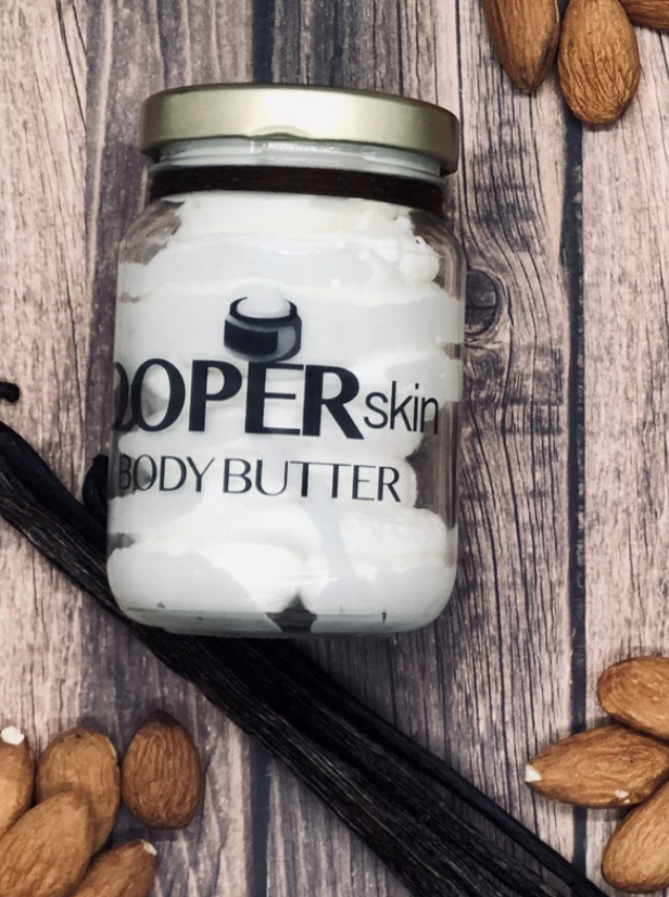 Clear jar of white body butter surrounded by piles of almonds on a wooden table