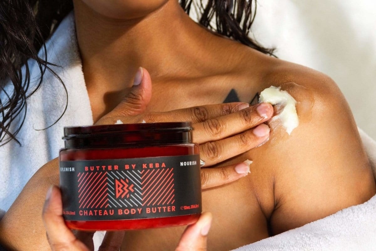 Model holding red and black jar of product and applying body butter to shoulder