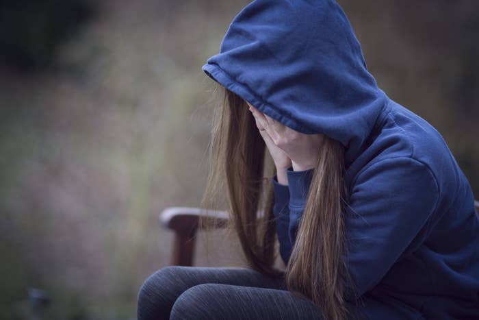 An anonymous teen wearing a hoodie, sitting in a chair, and covering her face with her hands