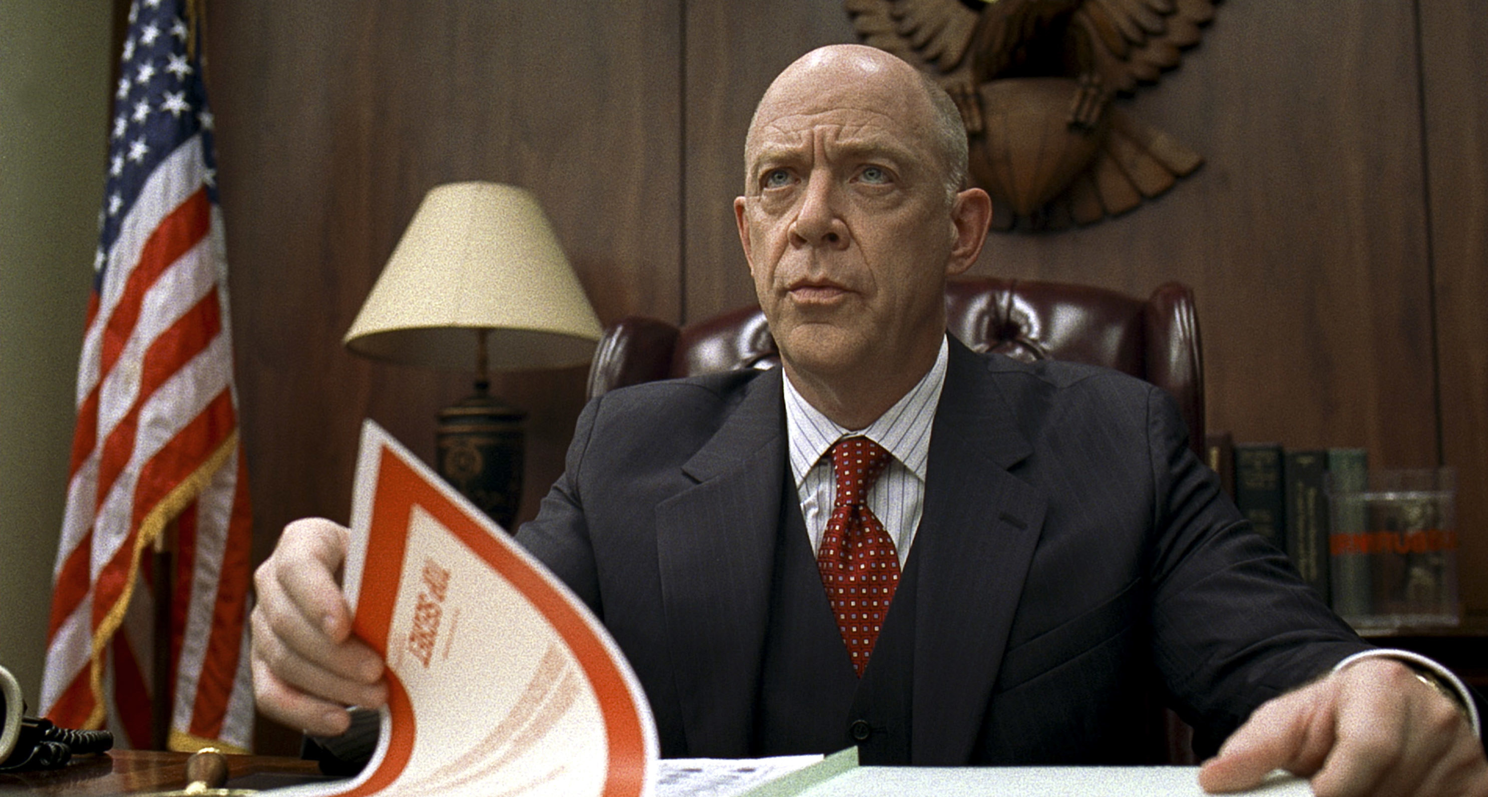 JK Simmons in a suit sitting in a large leather office chair in an offie looking serious