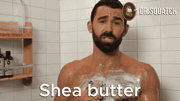 A man standing in a shower reading a bar of soap and saying, &quot;Shea butter&quot;