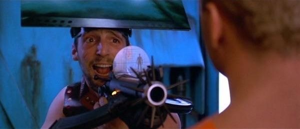 A man holding a futuristic gun to Bruce Willis and wearing a disguise on his head