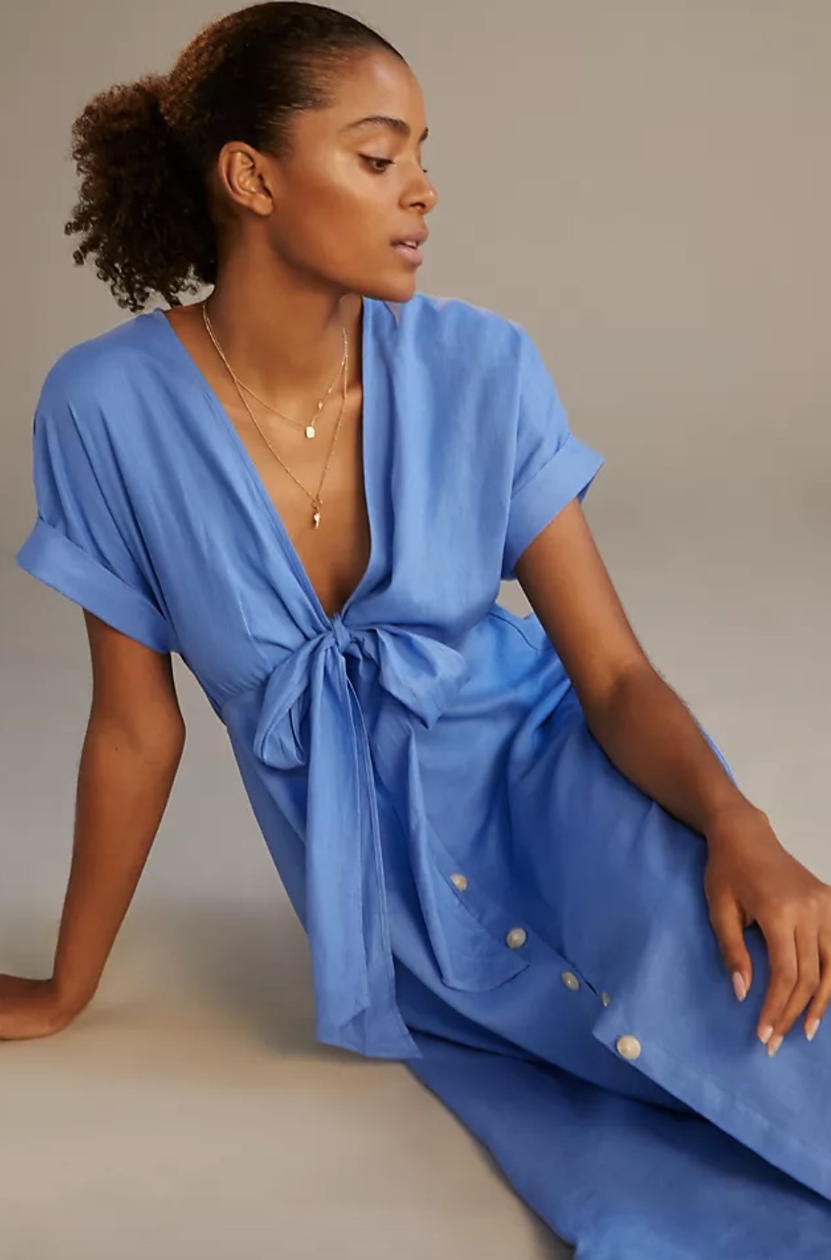 An adult sits and poses in the blue linen dress with a tie front and layer of buttons down the front