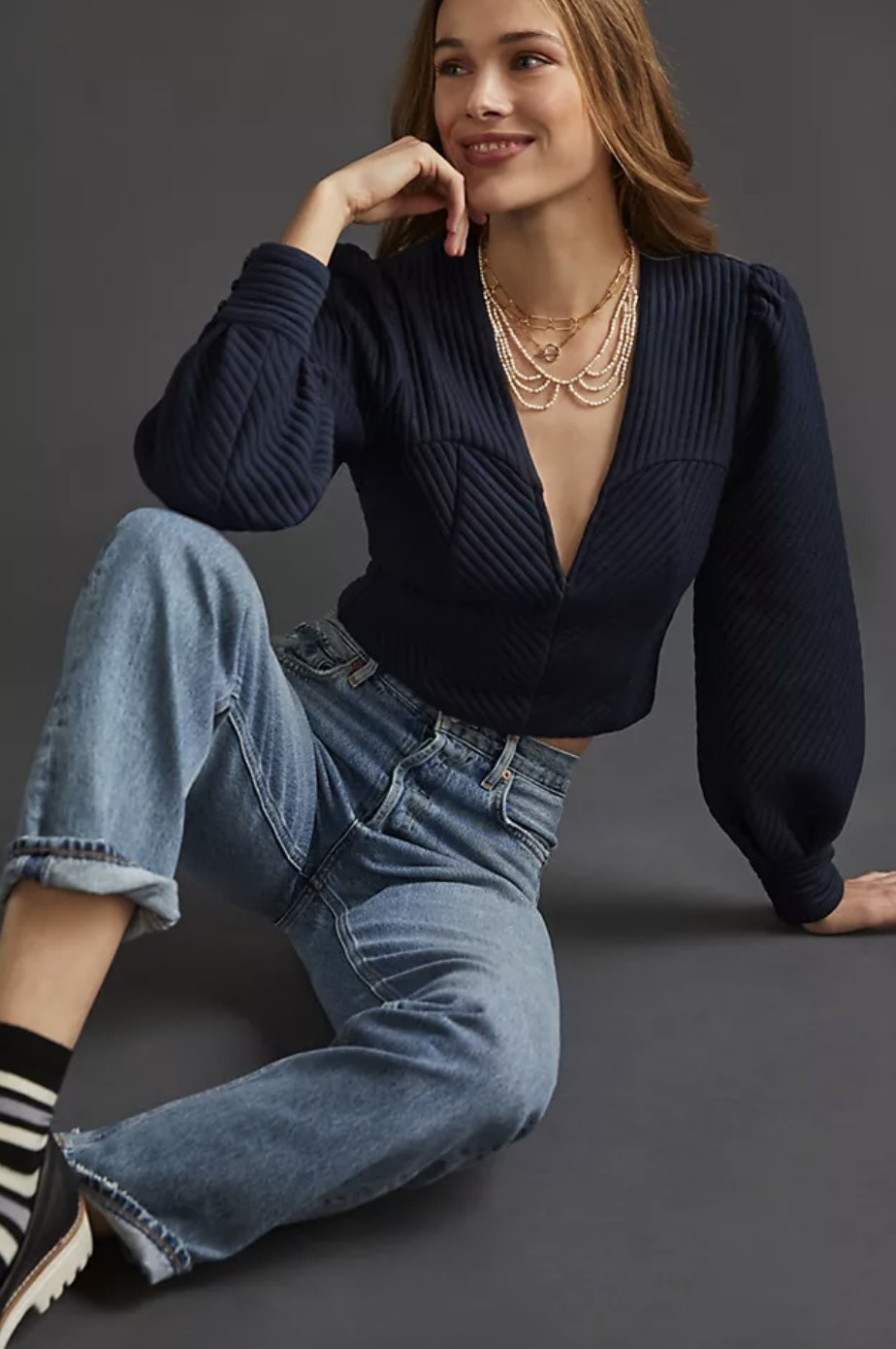 An adult wears a dark blue pullover with ribbed detailing paired with jeans a nd hold jewelry