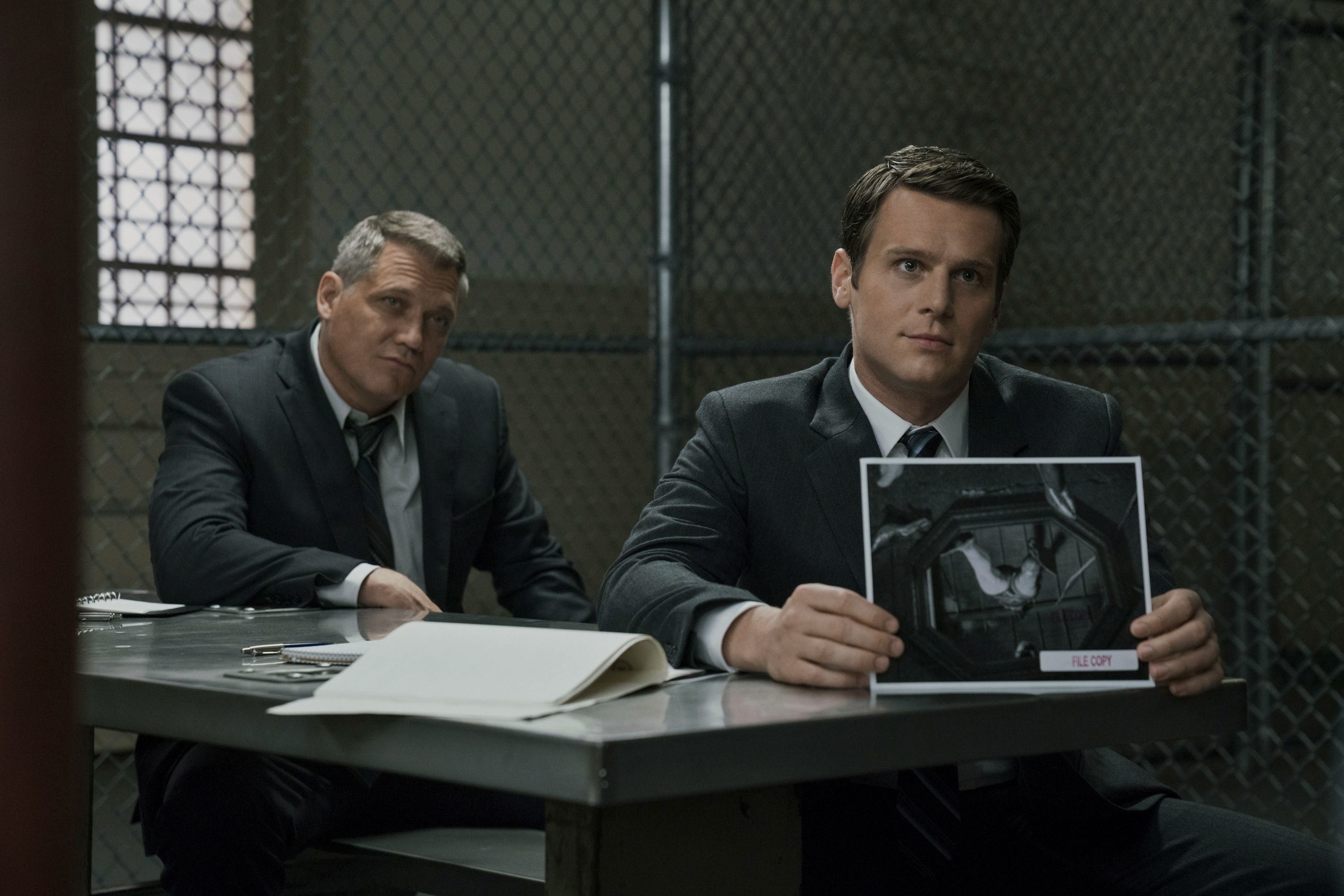 Holt McCallany as FBI agent Bill Tench sitting next to Jonathan Groff as agent Holden Ford presenting a picture of a dead body to someone in a prison in &quot;Mindhunter&quot;