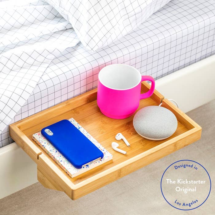 the wooden floating nightstand attached to a bed and holding a phone, mug, earbuds, and speaker