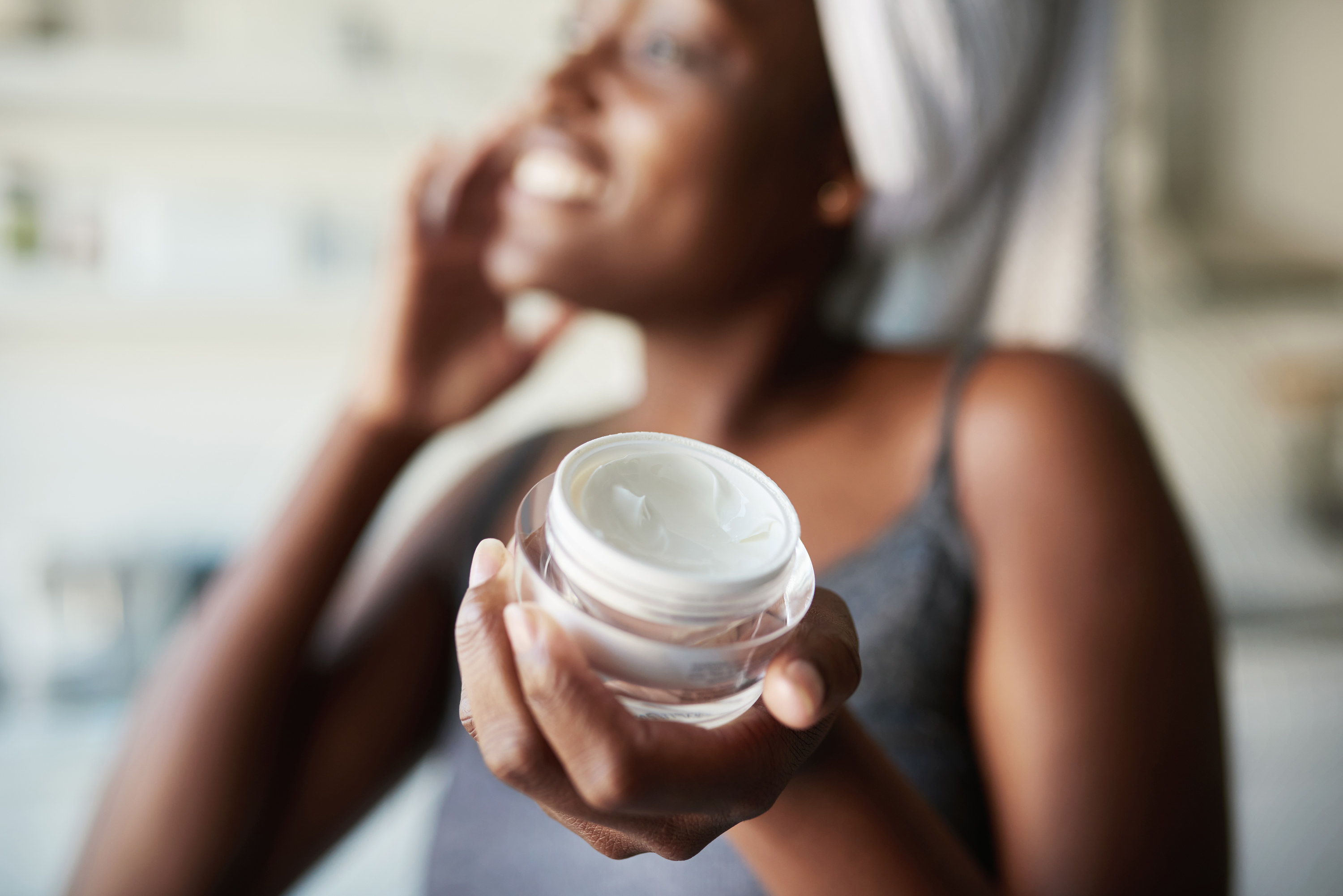 A woman holding a jar of moisturizer and is applying it in the background