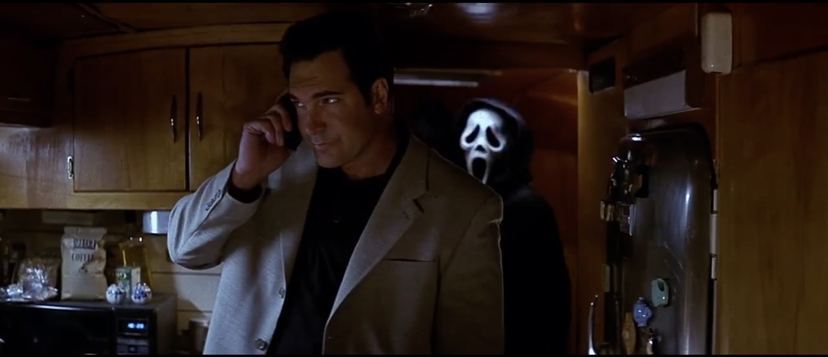 Patrick Warburton talks on the phone as Ghostface approaches him from behind