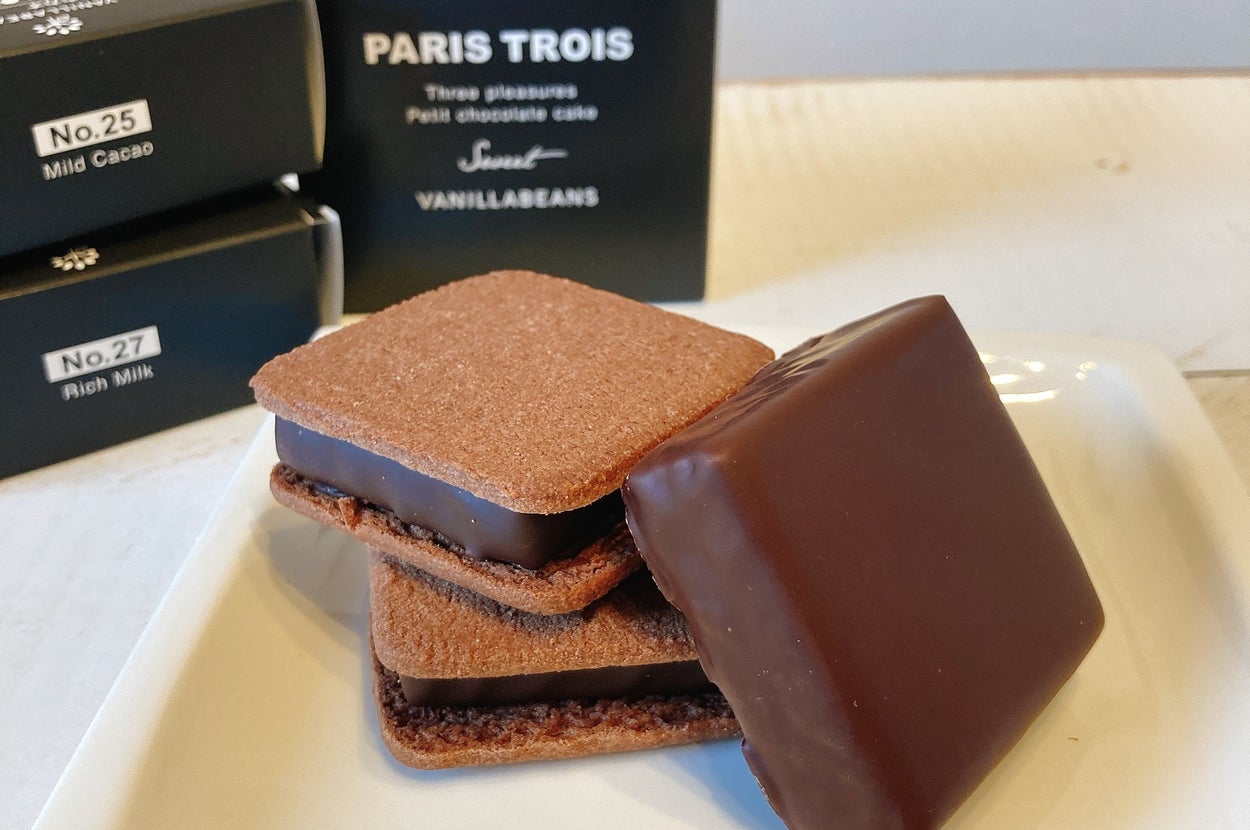 チョコレートを挟んだスクエアクッキーが皿に載っている。背景に「PARIS TROIS」などの黒いパッケージが見える。