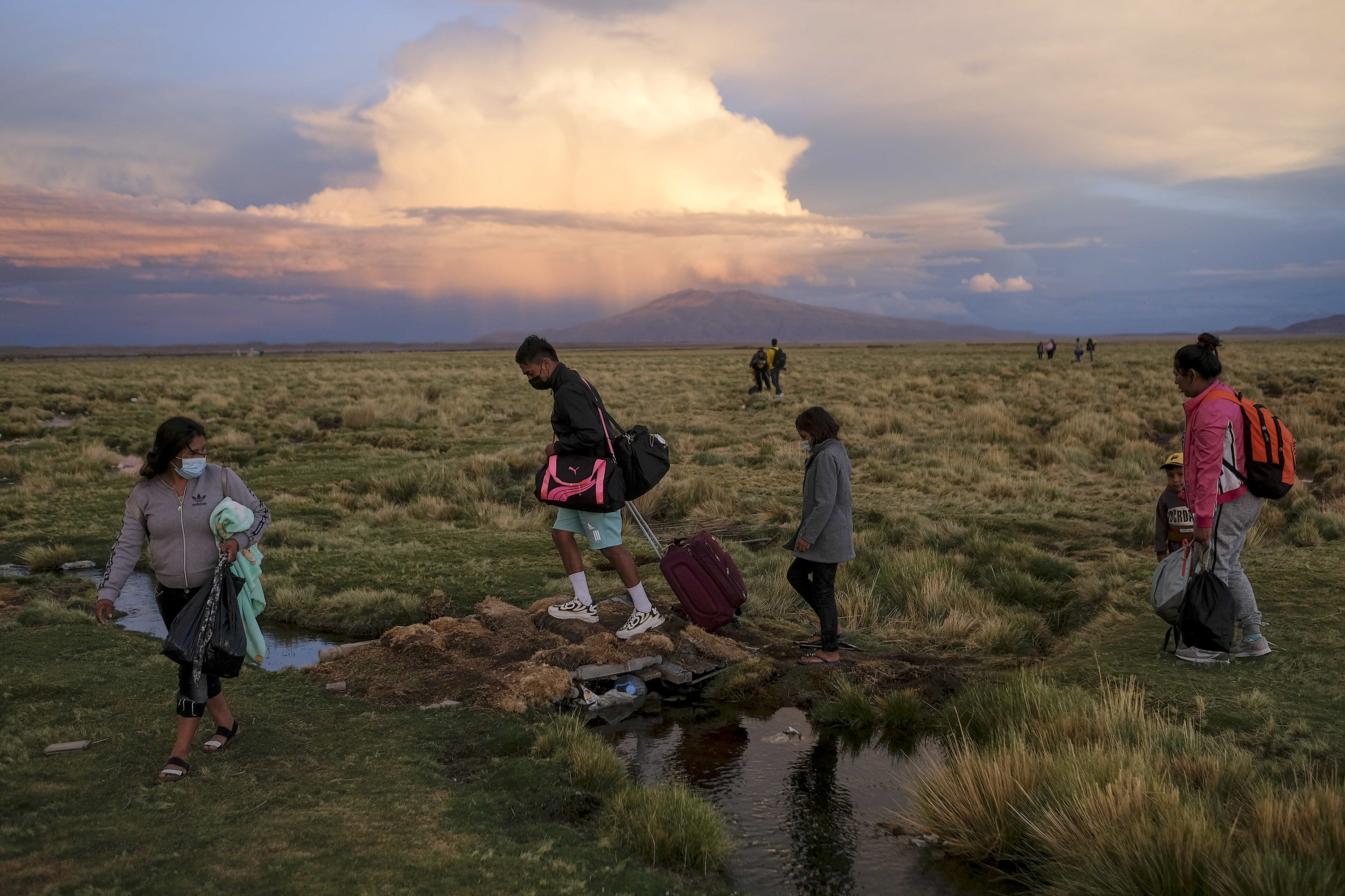People carrying luggage and other belongings walk past a small stream of water amid a barren landscape