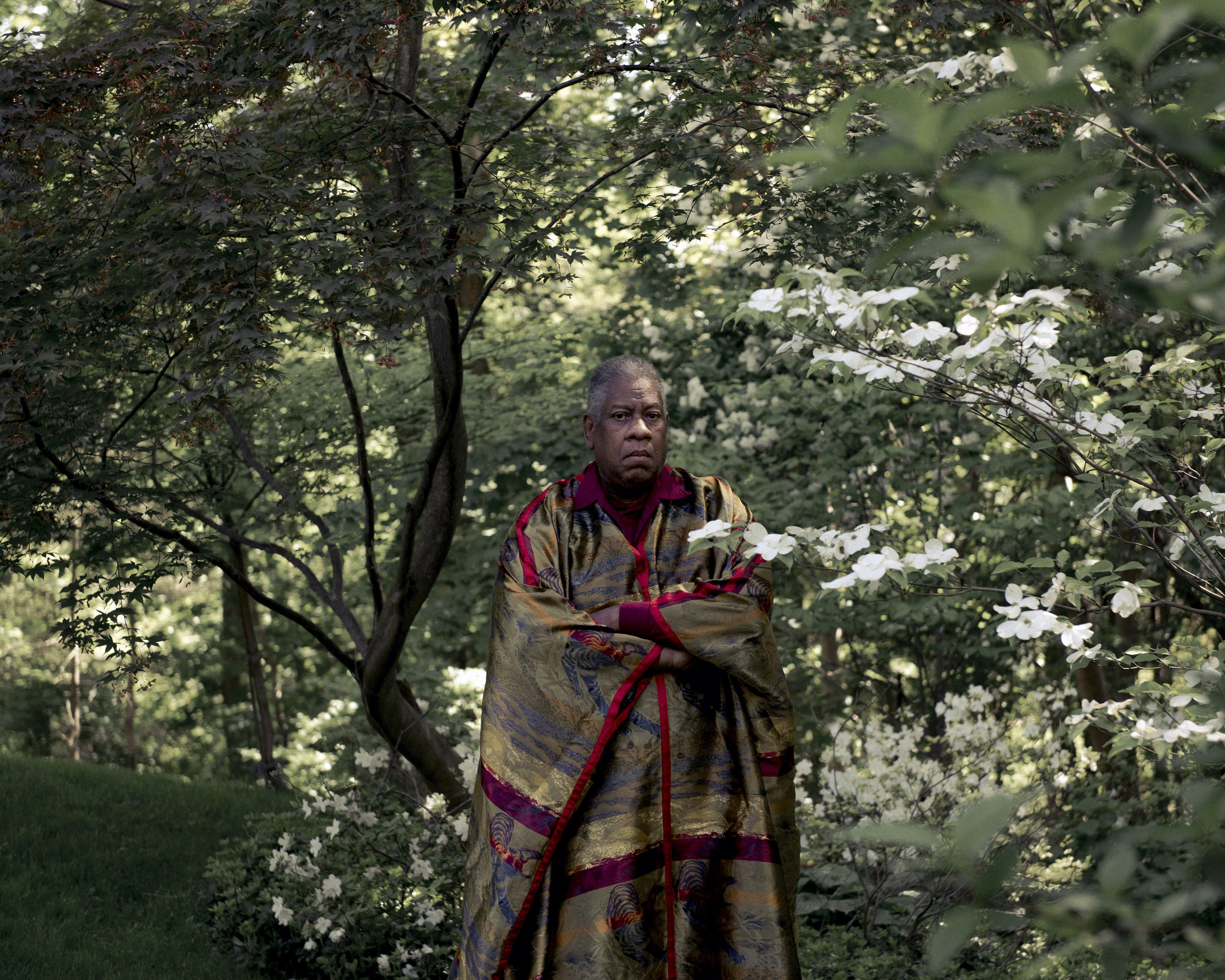 Talley, wearing a long, multicolored robe, stands with his arms crossed in front of many trees at his home in White Plains in 2018