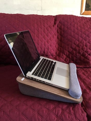 The wooden lap desk with a pillowed fabric base resting on a couch with a laptop elevated on it