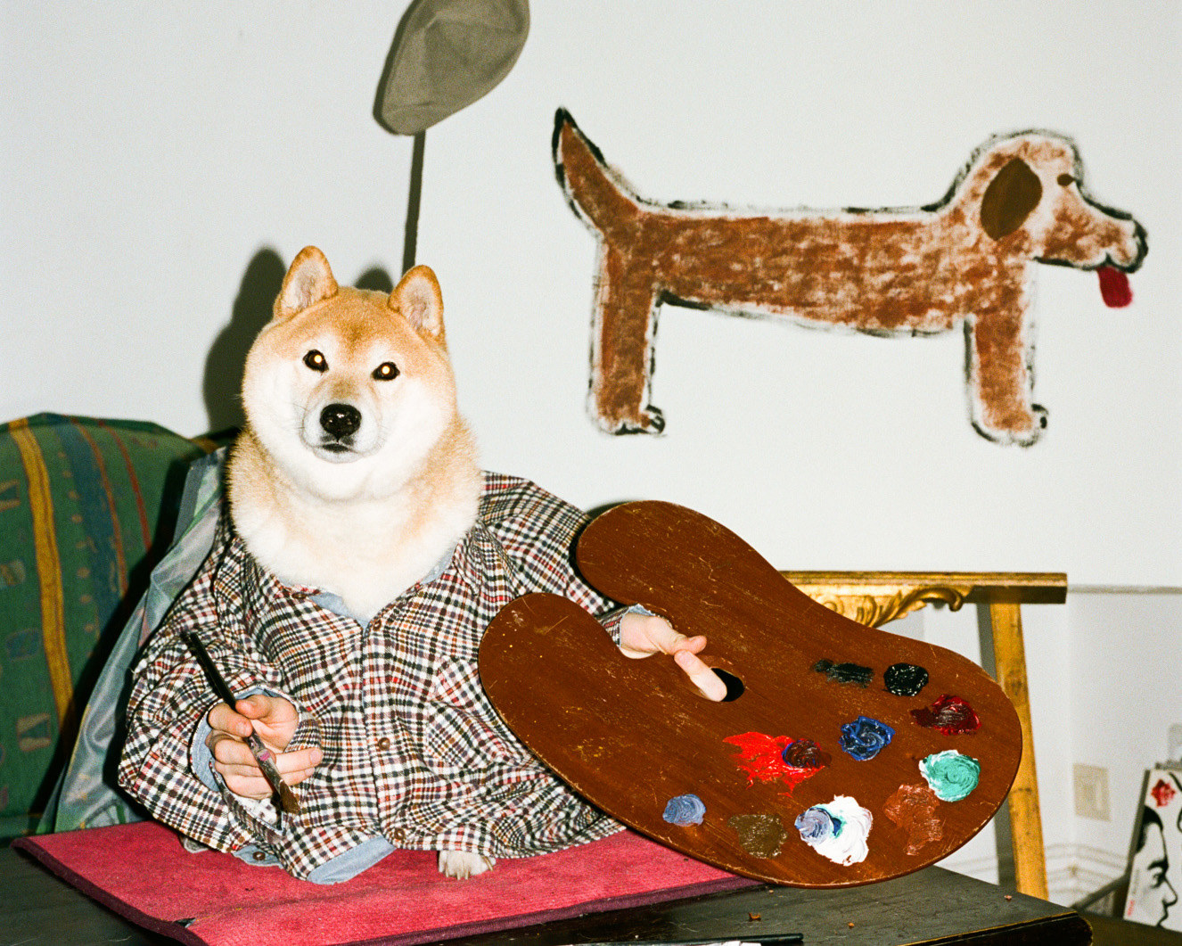 A dog seated on a chair appears to be holding a paint palette and a brush