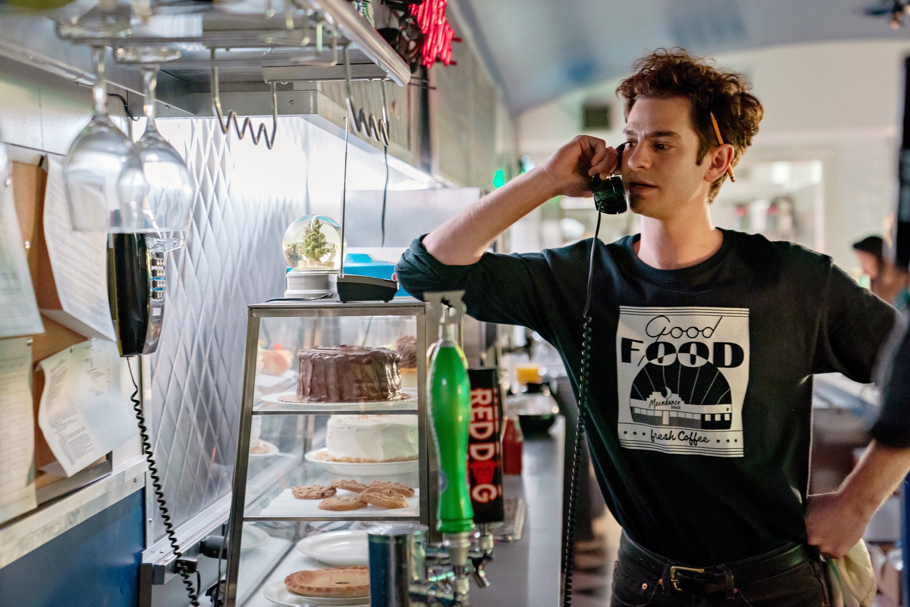 Andrew Garfield stands at a diner counter on the phone