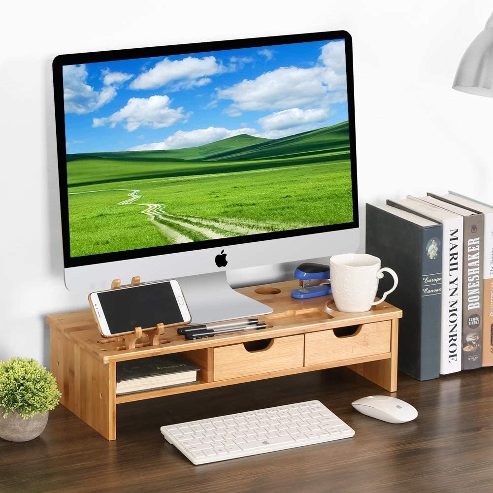 An Apple monitor sitting on a bamboo monitor stand with two drawers and various cutouts/storage for holding a mug, phone, stapler, and more
