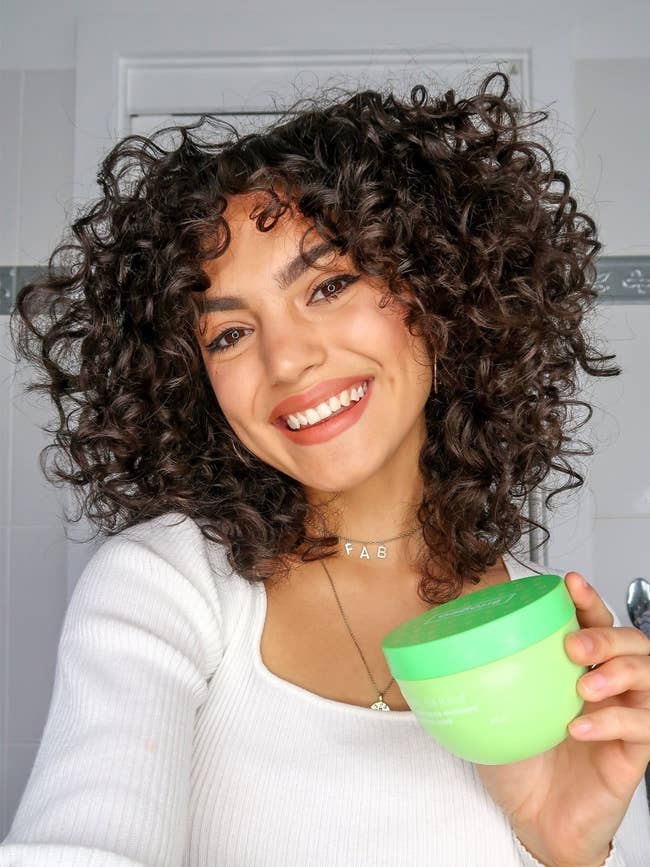 person with curly hair holding up a green jar of hair mask