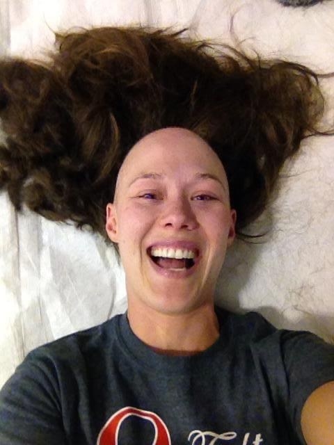 Cameron lying down and smiling as she shows off her bald head while a wig just above it