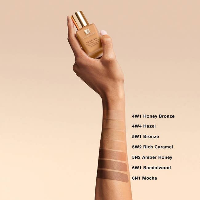 Model's hand holding a bottle of foundation with swatches of different foundation shades on their arm