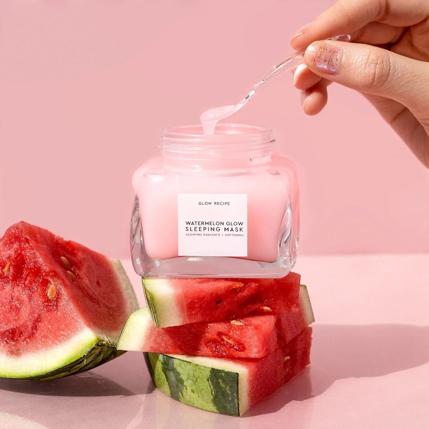 model&#x27;s hand dipping an applicator into a sleep mask jar, which is on top of sliced watermelon