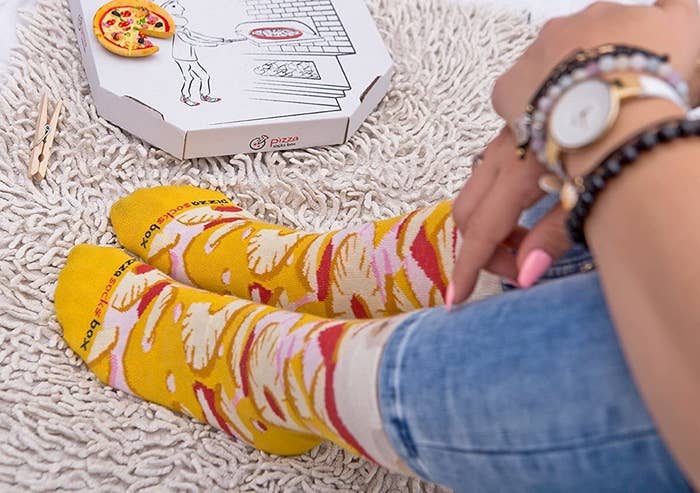 A person wearing a pair of pizza socks on a shaggy rug next to the box the socks come in