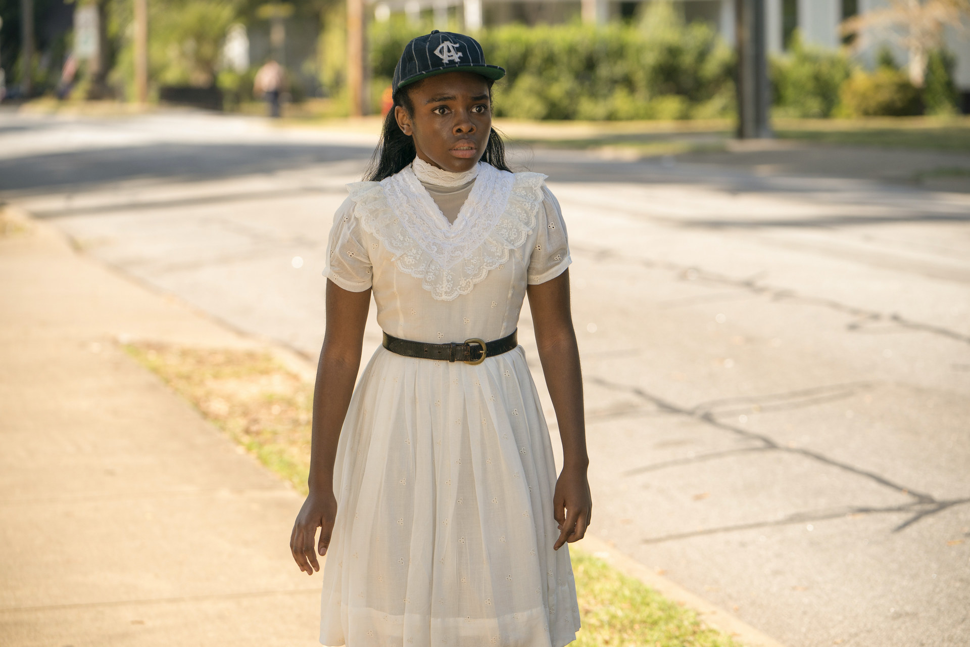 A Black teen girl walks along sidewalk in a white belted dress and baseball cap
