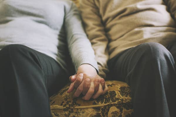 An older couple holds hands while sitting on the couch